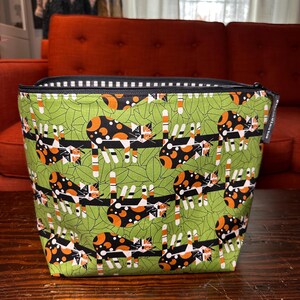 Puede incluir: Un neceser rectangular con cremallera y un estampado caprichoso de gatos. La bolsa presenta un fondo verde con ilustraciones de gatos negros, naranjas y blancos. La cremallera es negra con un ribete a rayas blancas y negras. La bolsa est&aacute; sobre una superficie de madera.