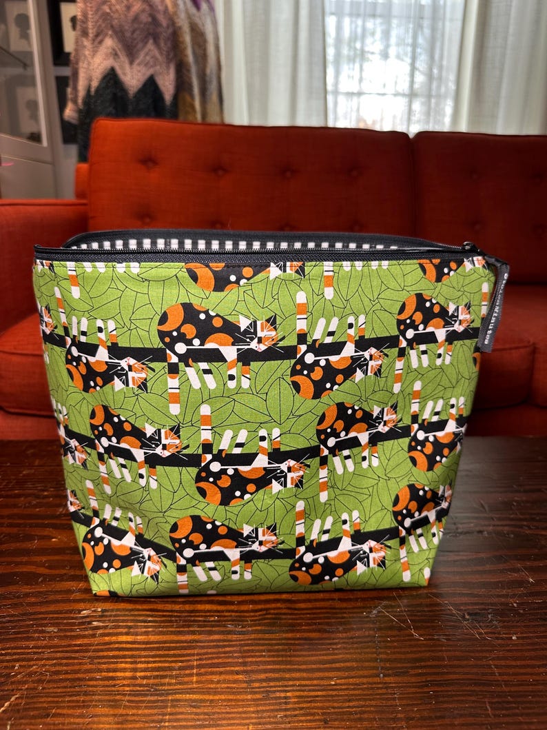 Puede incluir: Un neceser rectangular con cremallera y un estampado caprichoso de gatos. La bolsa presenta un fondo verde con ilustraciones de gatos negros, naranjas y blancos. La cremallera es negra con un ribete a rayas blancas y negras. La bolsa est&aacute; sobre una superficie de madera.
