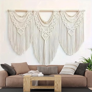 May include: A large, off-white macrame wall hanging with a wooden dowel. The macrame features intricate knotting patterns, beaded accents, and a fringed bottom. The wall hanging is displayed above a brown sofa with decorative pillows.
