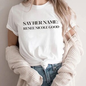 May include: White t-shirt with the text "SAY HER NAME: RENEE NICOLE GOOD" in black. The shirt is paired with blue jeans and a cream-colored cardigan.
