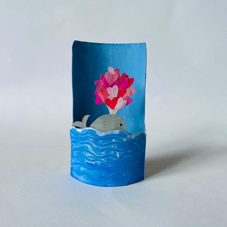 May include: A paper craft sculpture featuring a gray whale emerging from a textured blue painted ocean base. The whale is blowing a spray of various shades of pink and red paper hearts into the air against a light blue curved background.