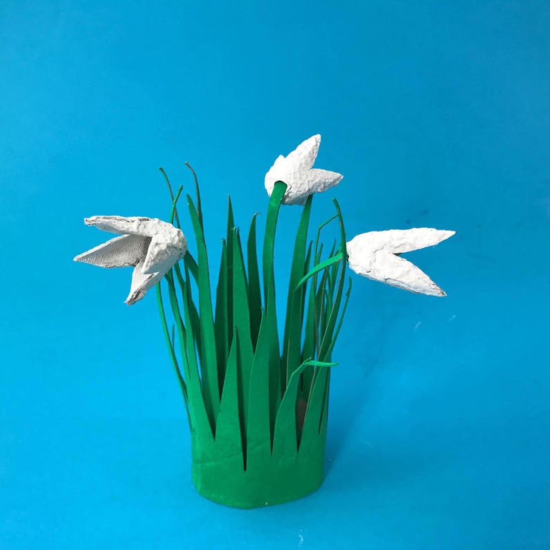 May include: A craft project featuring three white, textured, molded paper flowers attached to green stems. The stems are set within a cylindrical green base with jagged, grass-like cutouts at the top edge, creating a three-dimensional floral display.