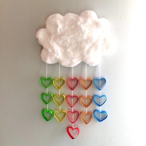 May include: A white fluffy cloud wall hanging with five vertical white strings suspended from it. Each string features a series of colorful paper heart loops in shades of green, yellow, red, orange, and blue, creating a rainbow effect.
