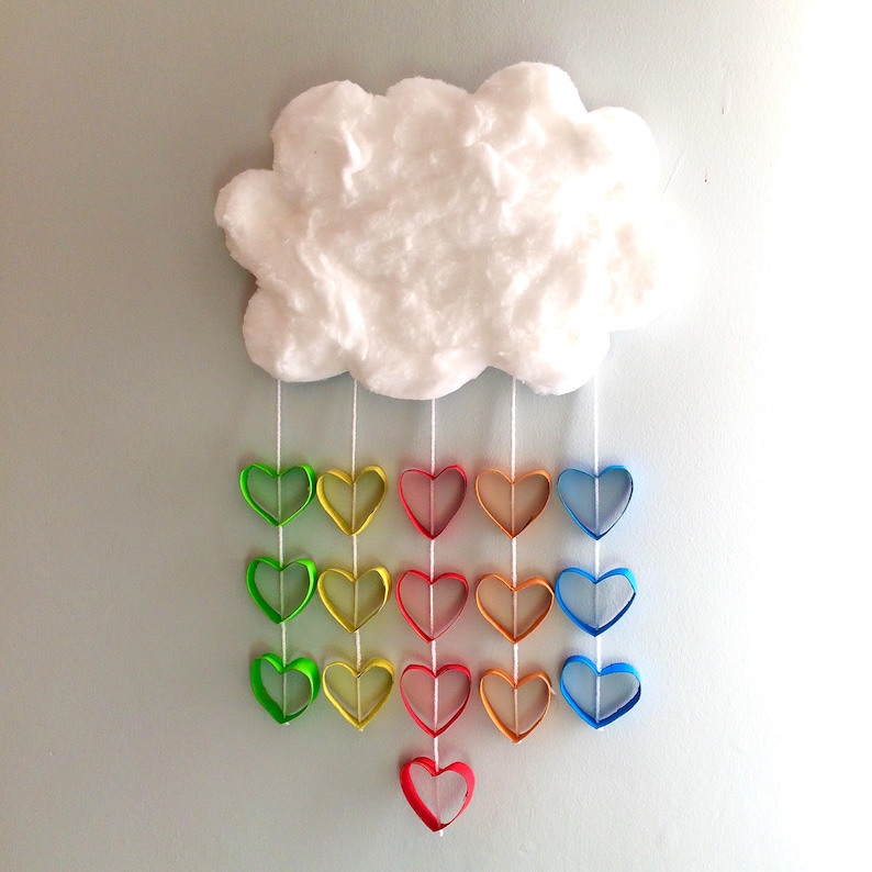 May include: A white fluffy cloud wall hanging with five vertical white strings suspended from it. Each string features a series of colorful paper heart loops in shades of green, yellow, red, orange, and blue, creating a rainbow effect.