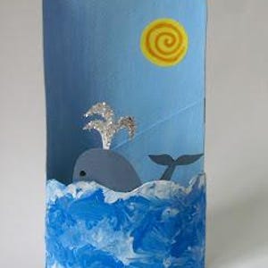 May include: A cylindrical craft project featuring a light blue background with a yellow spiral sun. A gray whale with a silver glitter spout emerges from a base painted with textured blue and white waves, creating a three-dimensional ocean scene.