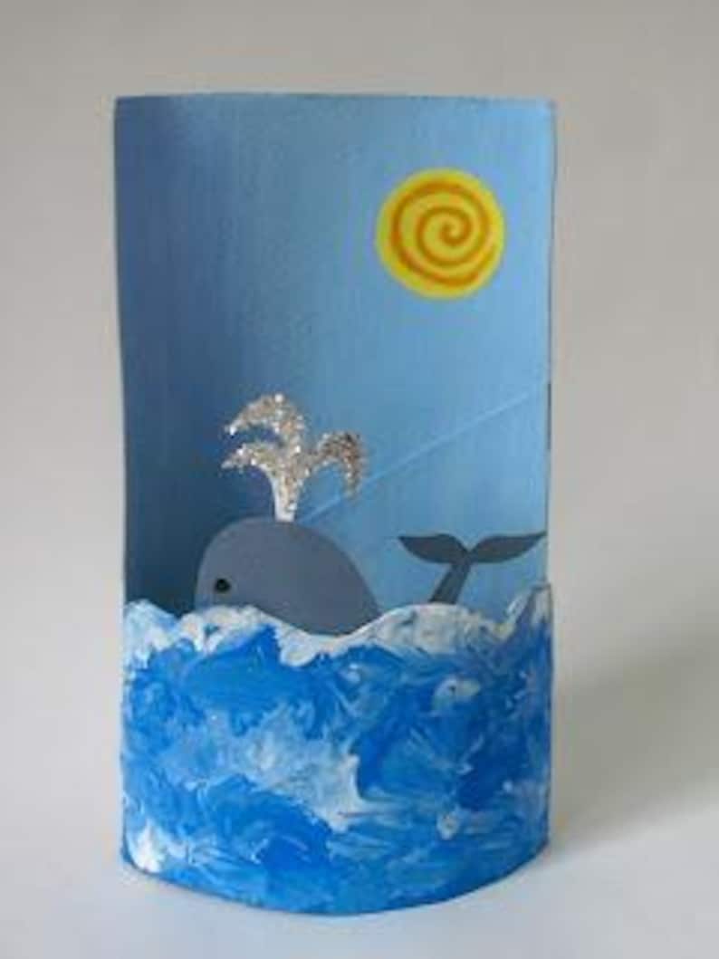 May include: A cylindrical craft project featuring a light blue background with a yellow spiral sun. A gray whale with a silver glitter spout emerges from a base painted with textured blue and white waves, creating a three-dimensional ocean scene.