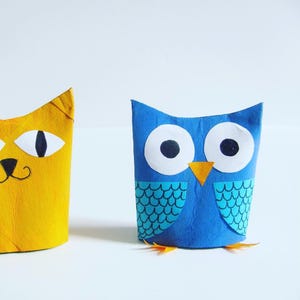 May include: A yellow cat figure with pointed ears and black facial features alongside a blue owl figure with large white eyes, a small orange beak, and dark blue wing patterns, both crafted from cylindrical paper tubes on a white surface.