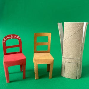May include: A red chair with white decorative scrollwork on the backrest, a plain yellow chair, and a cylindrical cardboard tube with a hand-drawn outline of a chair on its surface, all crafted from paper and cardboard materials.
