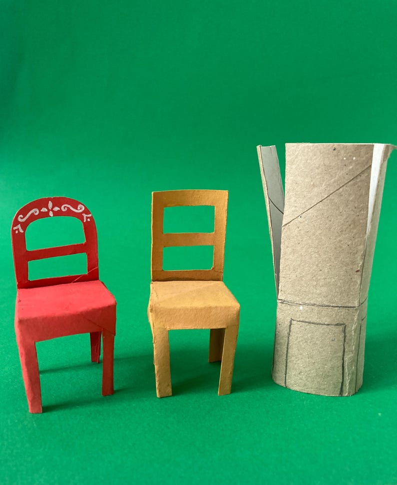 May include: A red chair with white decorative scrollwork on the backrest, a plain yellow chair, and a cylindrical cardboard tube with a hand-drawn outline of a chair on its surface, all crafted from paper and cardboard materials.