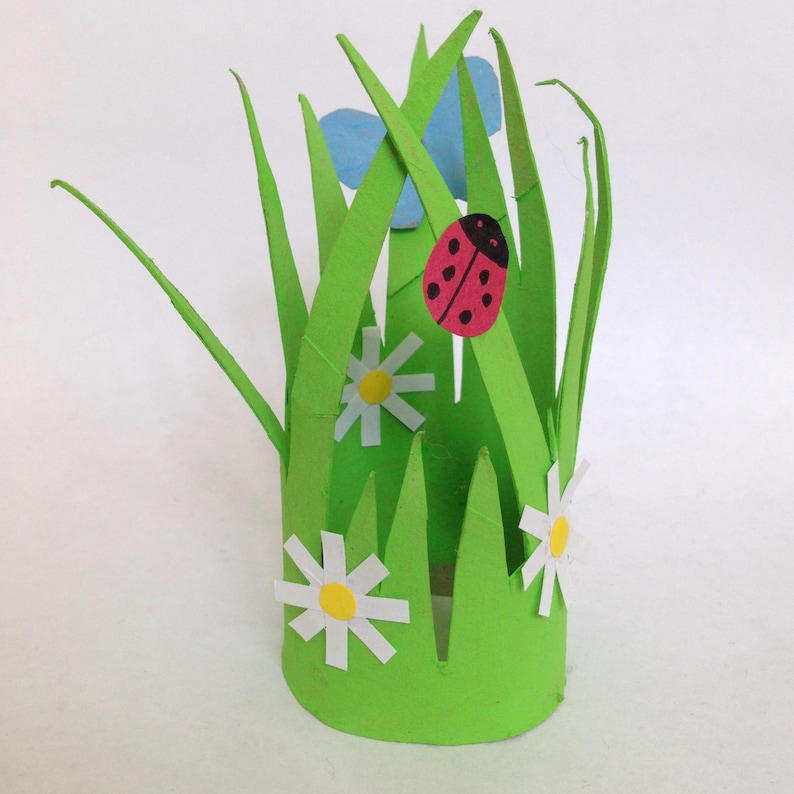 May include: A cylindrical green paper craft structure featuring tall, pointed grass blades. The exterior is decorated with two white daisies with yellow centers, a red ladybug with black spots, and a light blue butterfly cutout attached to the top.