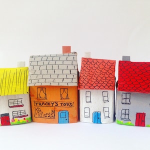 May include: Four miniature houses crafted from painted cardboard. The first has a yellow roof, the second is orange with the text TRACEY'S TOYS, the third is white with a red roof, and the fourth is gray with a red roof and black window details.