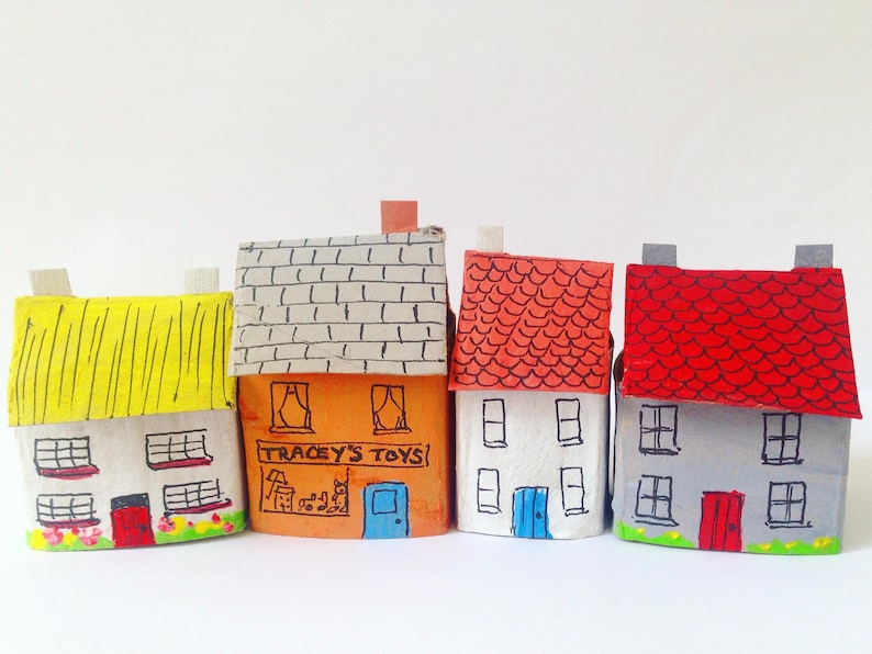 May include: Four miniature houses crafted from painted cardboard. The first has a yellow roof, the second is orange with the text TRACEY'S TOYS, the third is white with a red roof, and the fourth is gray with a red roof and black window details.