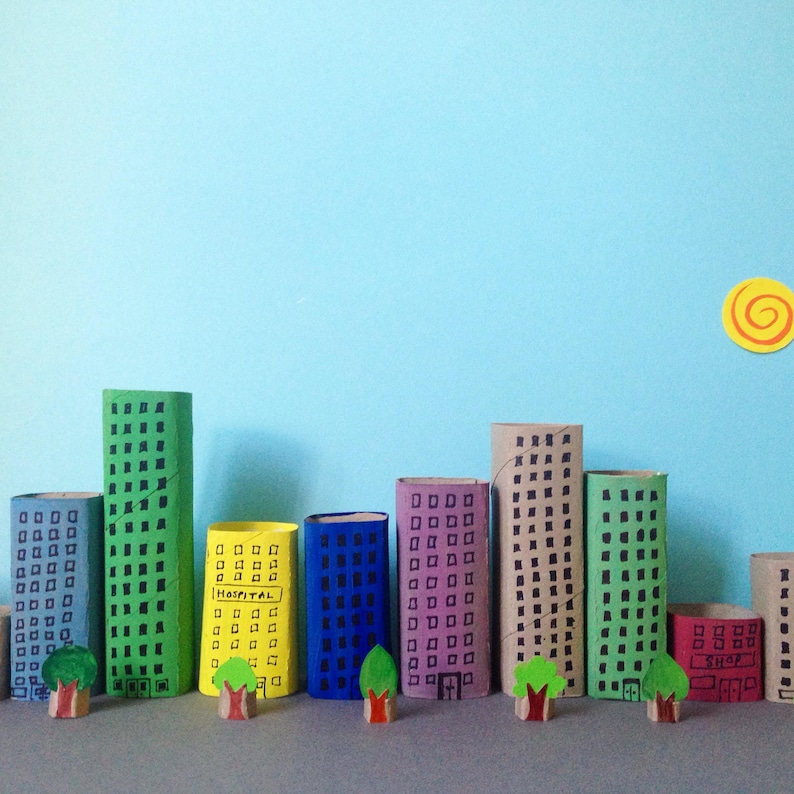 May include: A miniature city skyline crafted from cardboard tubes painted in various colors including green, yellow, blue, purple, and red. Each tube features hand-drawn black windows and doors, with small paper trees and a yellow sun cutout.