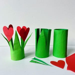 May include: A craft project featuring three green cardboard tubes. One tube is decorated with red paper hearts as flowers and green paper leaves. Another tube is partially cut into leaf shapes, and the third is a plain green tube with loose paper hearts.