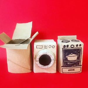 May include: Three miniature appliances crafted from brown cardboard. On the left is an open box, in the center is a washing machine with a circular door, and on the right is a stove with a black ink drawing of a pot inside the oven door.