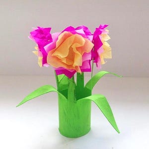 May include: A craft project featuring a bright green cylindrical base with attached green paper leaves. The top consists of several layered tissue paper flowers in vibrant shades of magenta and soft peach, arranged to resemble a small bouquet.