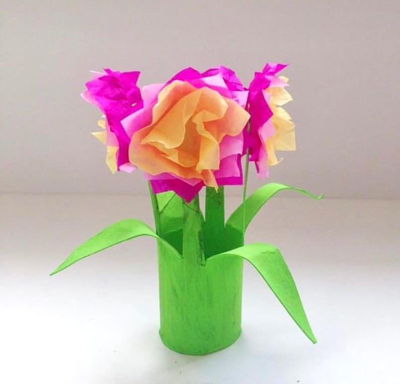 May include: A craft project featuring a bright green cylindrical base with attached green paper leaves. The top consists of several layered tissue paper flowers in vibrant shades of magenta and soft peach, arranged to resemble a small bouquet.