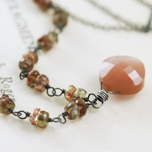 May include: A delicate necklace with a large peach-colored gemstone pendant. The necklace is made with small brown and green stones and silver wire.