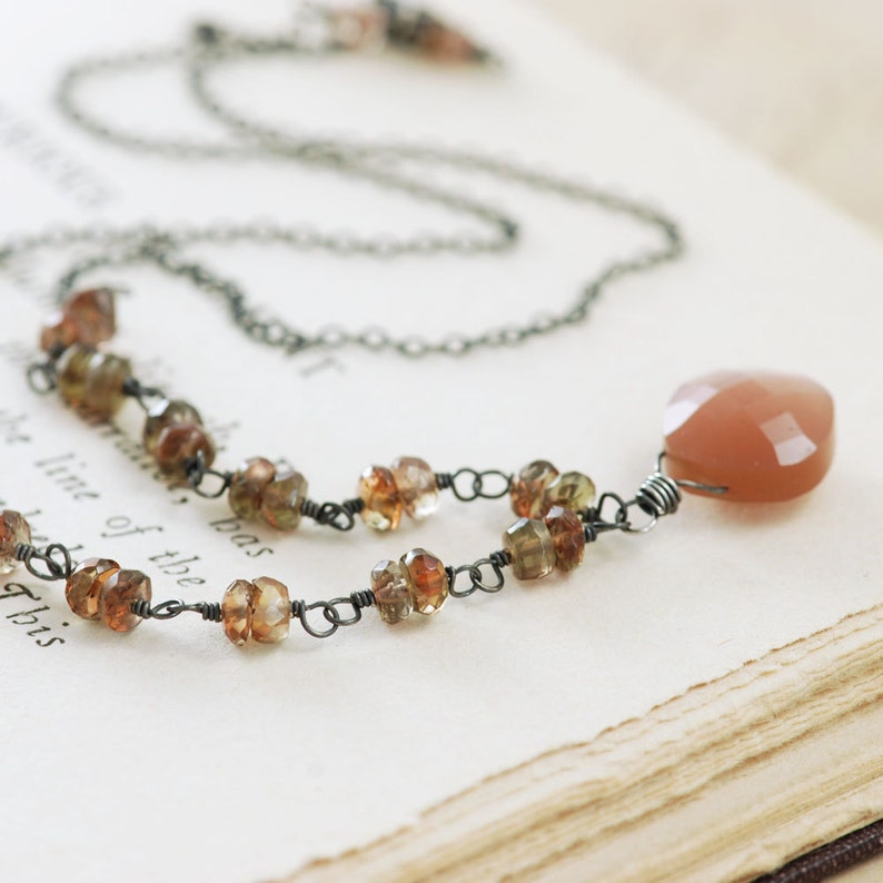 May include: A delicate necklace with a large peach-colored gemstone pendant. The necklace is made with a silver chain and small brown and green gemstones.