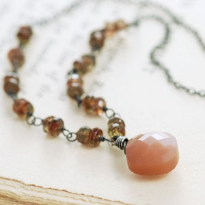 May include: A delicate necklace with a large peach-colored sunstone pendant. The necklace is made with small brown and green beads and a silver chain.