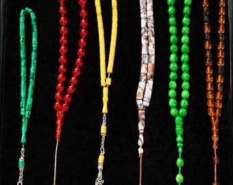 May include: Six prayer bead strands arranged vertically. The set includes green, red, yellow, marbled white and brown, bright green, and translucent amber beads. Each strand features a decorative metal tassel or a single bead at the bottom end.