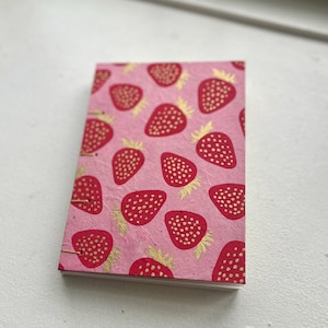 May include: A small, rectangular notebook with a pink cover featuring a pattern of red strawberries with gold accents. The notebook is bound with gold-coloured wire and has a textured paper appearance.
