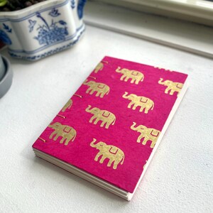 May include: A small, rectangular notebook with a vibrant pink cover. The cover features a repeating pattern of gold elephant illustrations. The notebook has white pages, and the binding is visible along the spine.