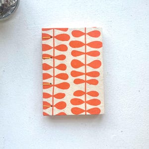 May include: A rectangular notebook with a cream-colored cover featuring a repeating orange leaf-stem pattern. The spine is exposed and bound with visible yellow thread stitching, securing the pages together in a simple, functional design.