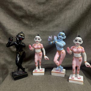 May include: Four small statues of figures in various poses. One is black, the others are pink and blue. The figures are standing on white rectangular bases. The background is a textured, gray fabric.
