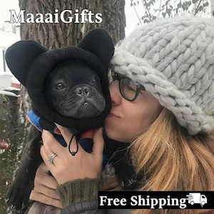 Könnte beinhalten: Ein schwarzer Französisch-Bulldoggen-Welpe trägt eine schwarze Kapuzenjacke mit Bärenohren. Der Welpe wird von einer Person gehalten, die eine graue Strickmütze und eine Brille trägt. Der Text "MaaaiGifts" befindet sich in der oberen linken Ecke.