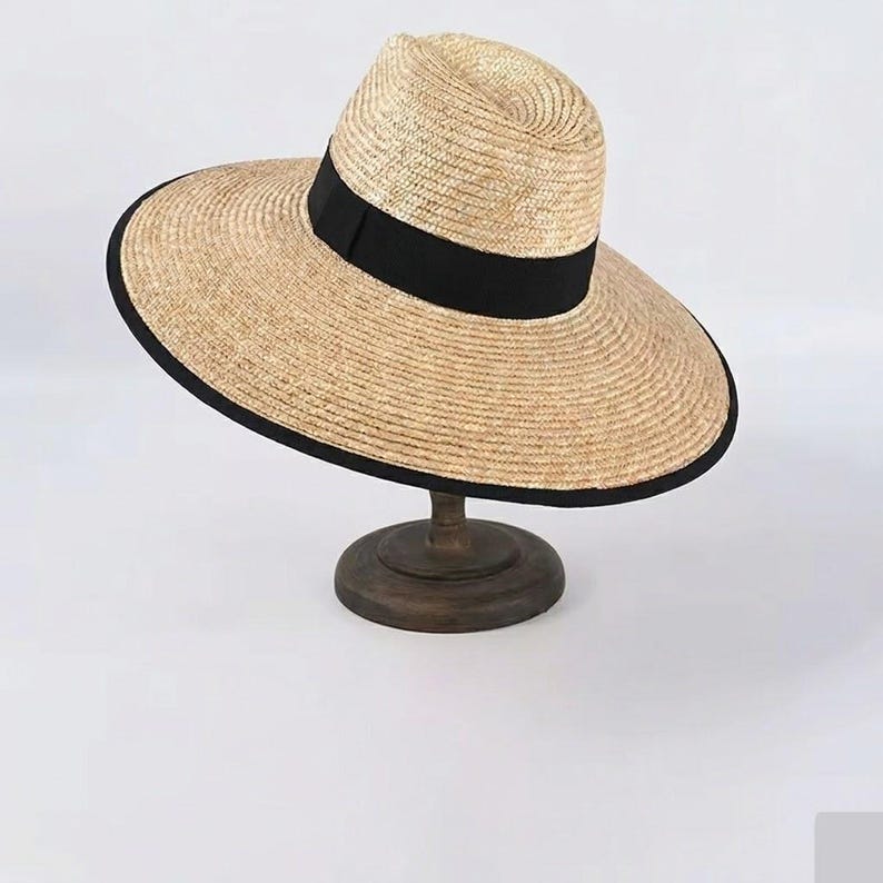 May include: A wide-brimmed straw hat with a black band and trim. The hat is displayed on a wooden stand. The hat's design is classic and suitable for sunny weather. The brim measures approximately 30 cm.