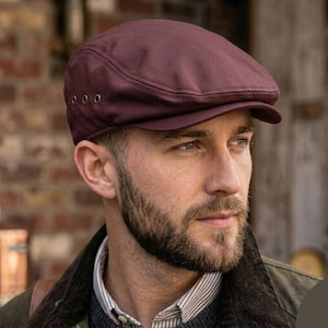 May include: A maroon flat cap with a rounded crown and a small, stiff peak. The cap features three small metal eyelets on the side. This classic style hat is made of a smooth fabric and is often worn for a casual or semi-formal appearance.