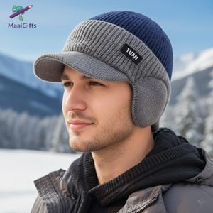 Puede incluir: Un gorro de invierno azul, gris y gris oscuro con orejeras y visera. El gorro tiene una textura de punto acanalado y una pequeña etiqueta rectangular negra que dice "YUAN". El gorro está diseñado para el clima frío.