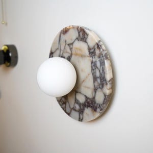 Pode incluir: Candeeiro de parede moderno com uma l&acirc;mpada esf&eacute;rica branca fixada a uma base oval com efeito m&aacute;rmore. O m&aacute;rmore tem um padr&atilde;o de veios brancos, bege e cinza escuro. O candeeiro est&aacute; montado numa parede branca.