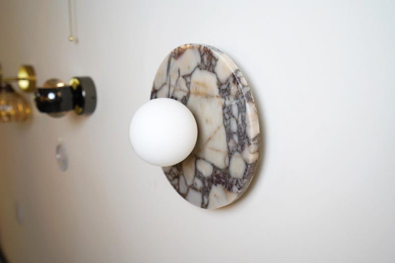 Pode incluir: Candeeiro de parede moderno com uma l&acirc;mpada esf&eacute;rica branca fixada a uma base oval com efeito m&aacute;rmore. O m&aacute;rmore tem um padr&atilde;o de veios brancos, bege e cinza escuro. O candeeiro est&aacute; montado numa parede branca.