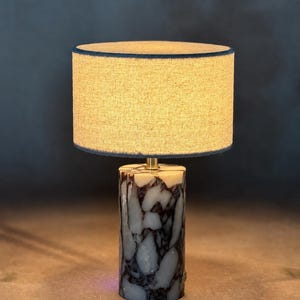 May include: A table lamp featuring a cylindrical base made of white and dark purple marbled stone. The lamp is topped with a round, textured beige fabric lampshade that emits a warm glow, supported by a small brass-colored metal connector.