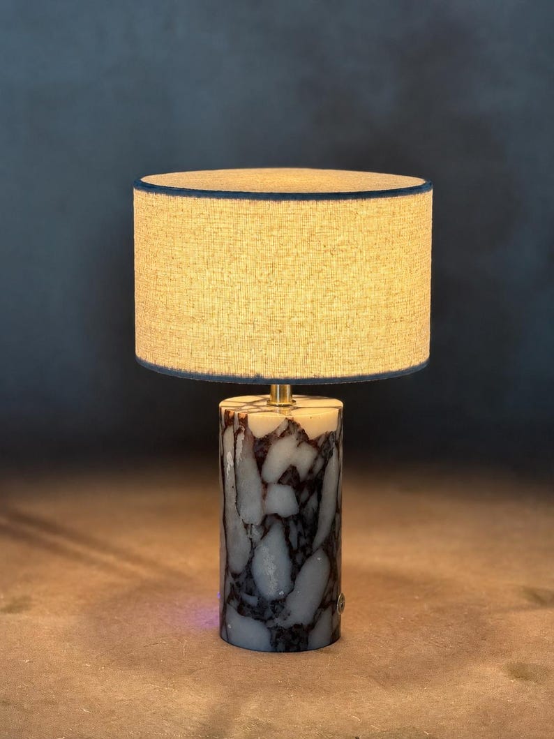 May include: A table lamp featuring a cylindrical base made of white and dark purple marbled stone. The lamp is topped with a round, textured beige fabric lampshade that emits a warm glow, supported by a small brass-colored metal connector.