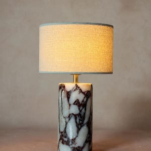 May include: A table lamp featuring a cylindrical base made of white and dark maroon veined marble. The lamp has a gold-toned metal neck and a cylindrical fabric lampshade in a neutral beige tone that emits a warm, soft glow when illuminated.