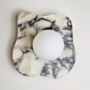 May include: A decorative object featuring a white sphere resting on a marble tray. The tray has a unique, organic shape with a cream and grey marbled pattern. The sphere is smooth and opaque, contrasting with the tray's textured surface.