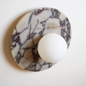 Pode incluir: Uma arandela decorativa com uma esfera branca adjacente a um oval de m&aacute;rmore. O m&aacute;rmore tem uma base branca com veios roxos escuros e cinzentos. A arandela &eacute; projetada para ilumina&ccedil;&atilde;o interior.