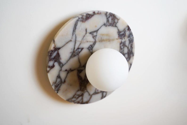 Pode incluir: Uma arandela decorativa com uma esfera branca adjacente a um oval de m&aacute;rmore. O m&aacute;rmore tem uma base branca com veios roxos escuros e cinzentos. A arandela &eacute; projetada para ilumina&ccedil;&atilde;o interior.