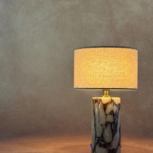 May include: A table lamp featuring a cylindrical base made of white and dark gray marbled stone. The lamp is topped with a drum-shaped lampshade in a textured beige fabric, which emits a warm glow. A small gold-toned metal connector joins the base.
