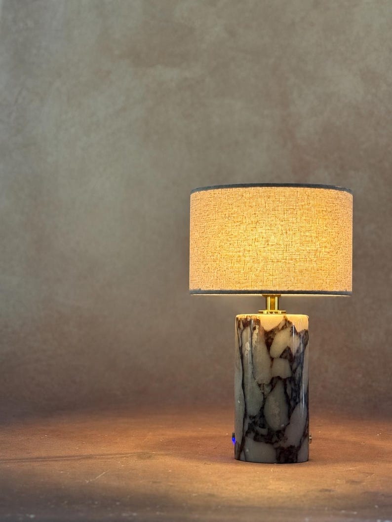 May include: A table lamp featuring a cylindrical base made of white and dark gray marbled stone. The lamp is topped with a drum-shaped lampshade in a textured beige fabric, which emits a warm glow. A small gold-toned metal connector joins the base.