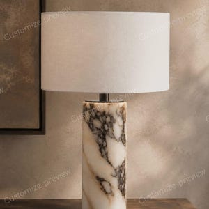 Puede incluir: Una lámpara de mesa con una base cilíndrica de piedra veteada en blanco y gris oscuro. La lámpara está rematada con una pantalla de tambor grande, lisa y de color blanco roto. La base tiene un acabado liso y pulido con vetas marcadas.