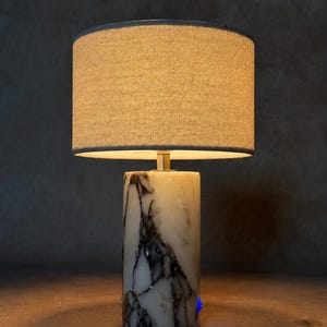 May include: A table lamp featuring a cylindrical base made of white marble with prominent dark grey and black veining. The lamp is topped with a circular, textured beige fabric drum shade that emits a warm glow from the light source within.