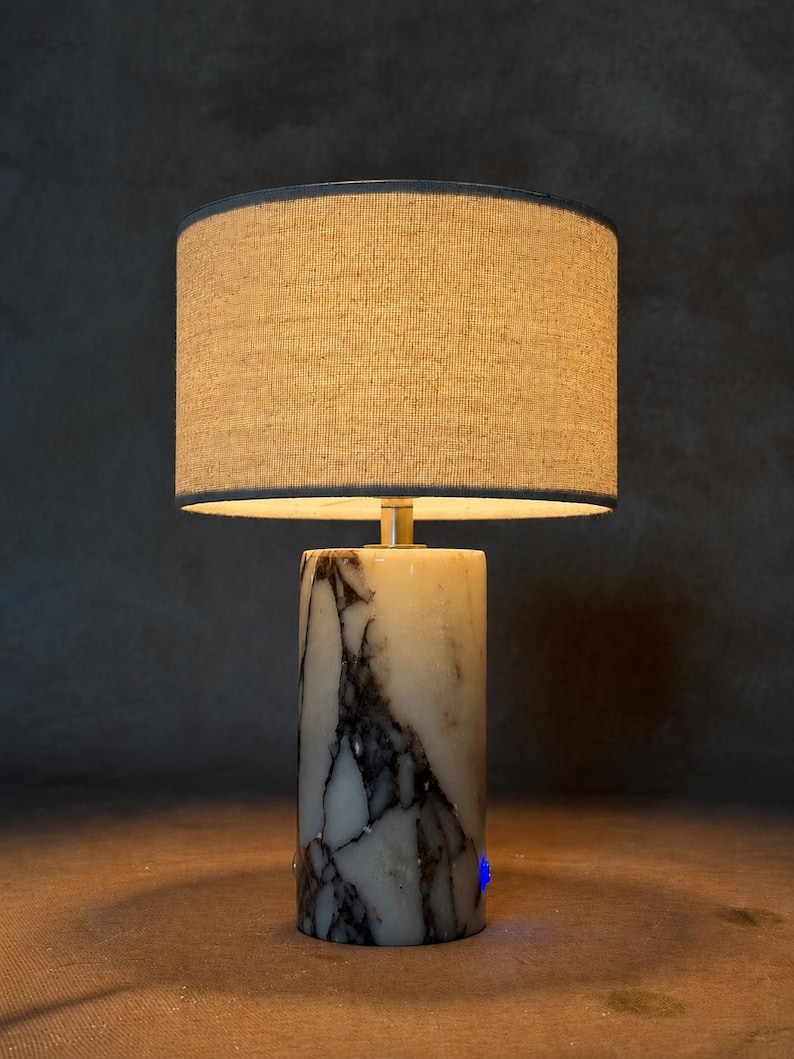 May include: A table lamp featuring a cylindrical base made of white marble with prominent dark grey and black veining. The lamp is topped with a circular, textured beige fabric drum shade that emits a warm glow from the light source within.