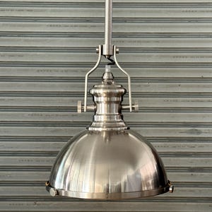 May include: Brushed nickel pendant light with a dome-shaped shade. The light fixture has a cylindrical stem and decorative hardware. The shade is a smooth, metallic silver colour. The light is suspended against a grey, horizontally-lined background.