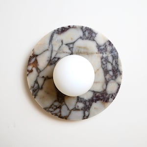 Puede incluir: Un posavasos redondo de mármol con una esfera blanca en el centro. El posavasos tiene una mezcla de vetas blancas, grises y marrones. La esfera es de color blanco liso. Los artículos están sobre un fondo blanco.