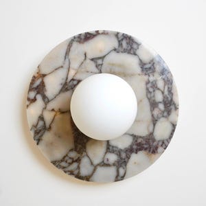 Pode incluir: Uma base redonda de m&aacute;rmore com uma esfera branca no centro. A base tem um padr&atilde;o natural de branco, castanho e cinza. A esfera &eacute; lisa e opaca. O design &eacute; simples e moderno.
