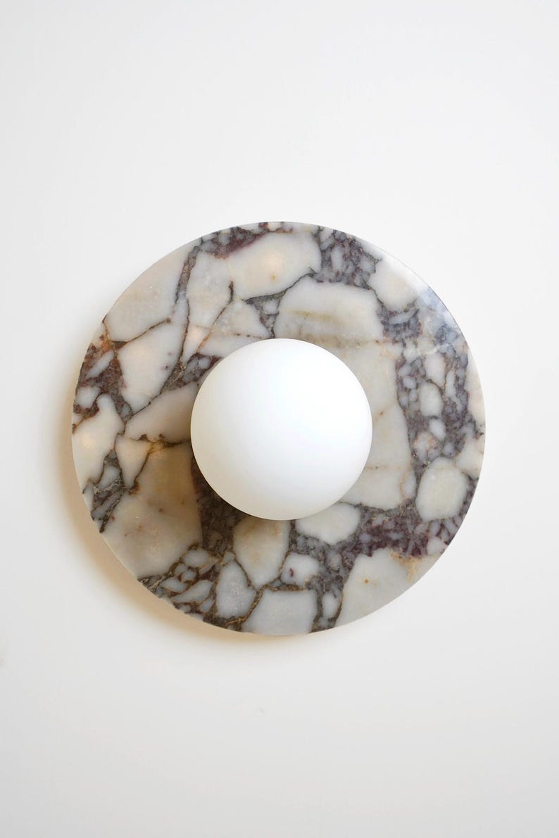 Pode incluir: Uma base redonda de m&aacute;rmore com uma esfera branca no centro. A base tem um padr&atilde;o natural de branco, castanho e cinza. A esfera &eacute; lisa e opaca. O design &eacute; simples e moderno.